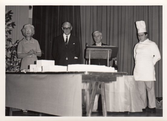 Black and white photograph of the celebration cake including headmaster and chef. | Unknown